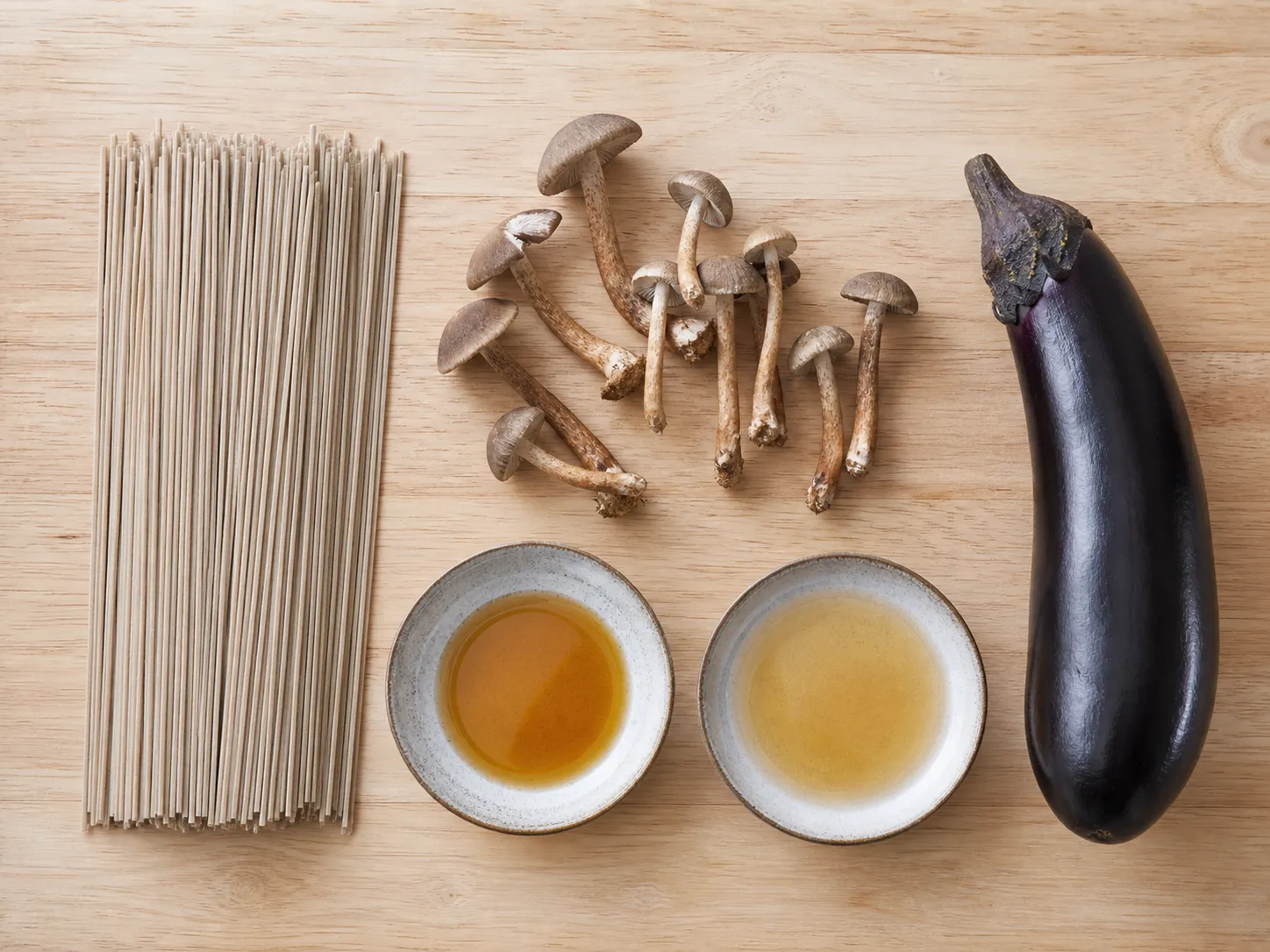 Flat lay of chitake soba ingredients: raw soba noodles, wild chitake mushrooms, a whole eggplant, sesame oil and dashi