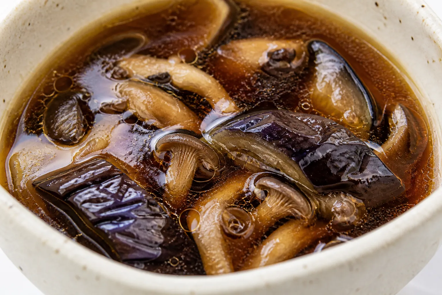 Close-up of chitake soba dipping broth with softened eggplant slices and mushroom pieces, oil beading on the surface