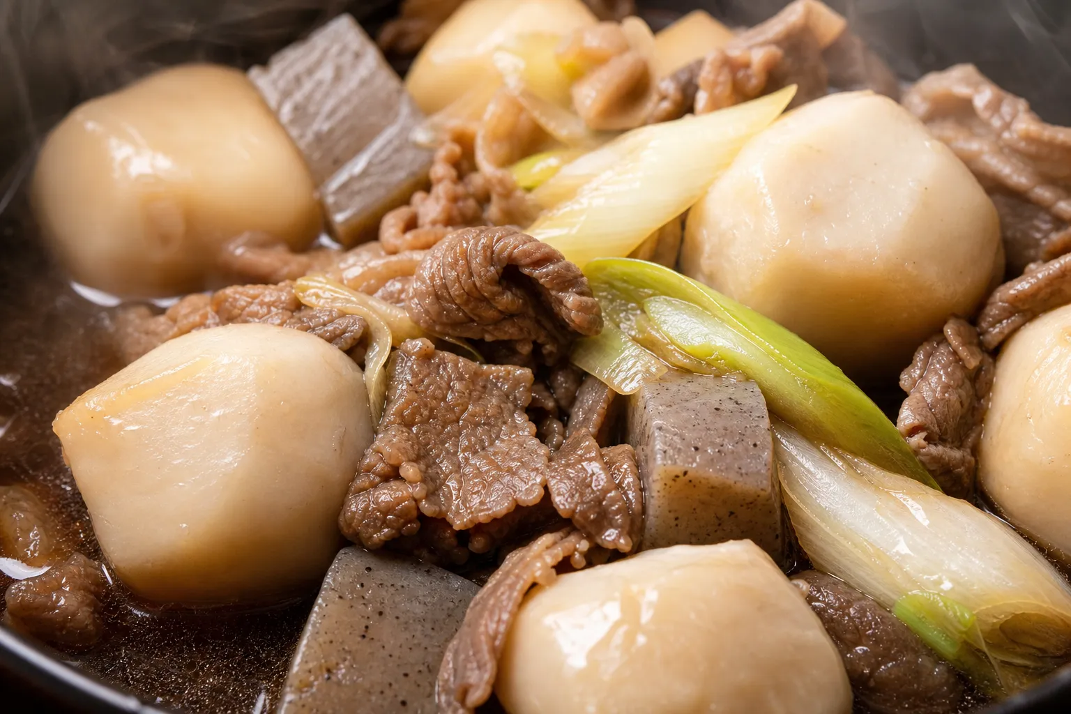 Close-up of imoni: soft taro, beef, and konnyaku in glossy soy broth