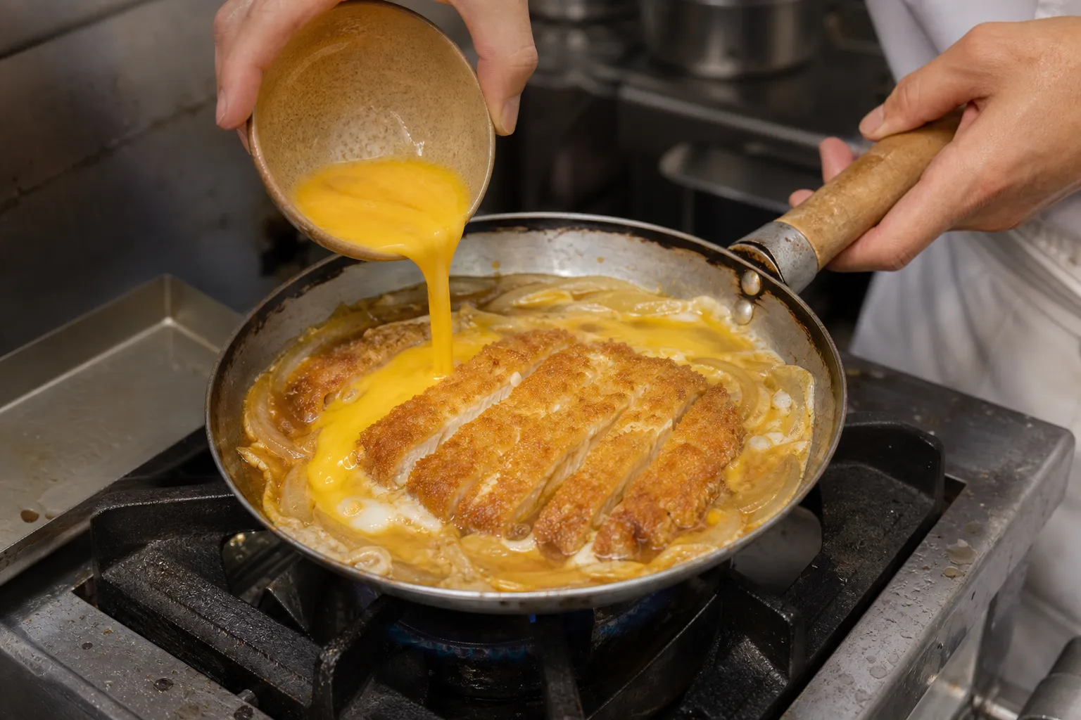 A cook pouring beaten egg over sliced tonkatsu and onions simmering in dashi-soy broth in a shallow pan