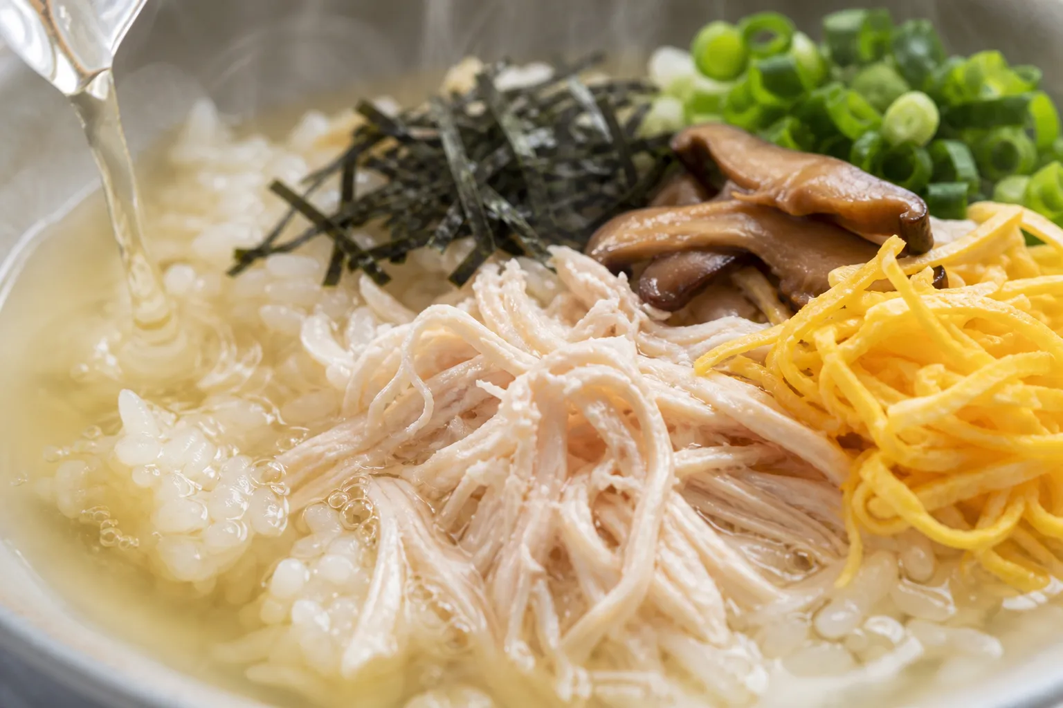 Close-up of keihan with shredded chicken, golden egg and nori as clear broth is poured over the rice