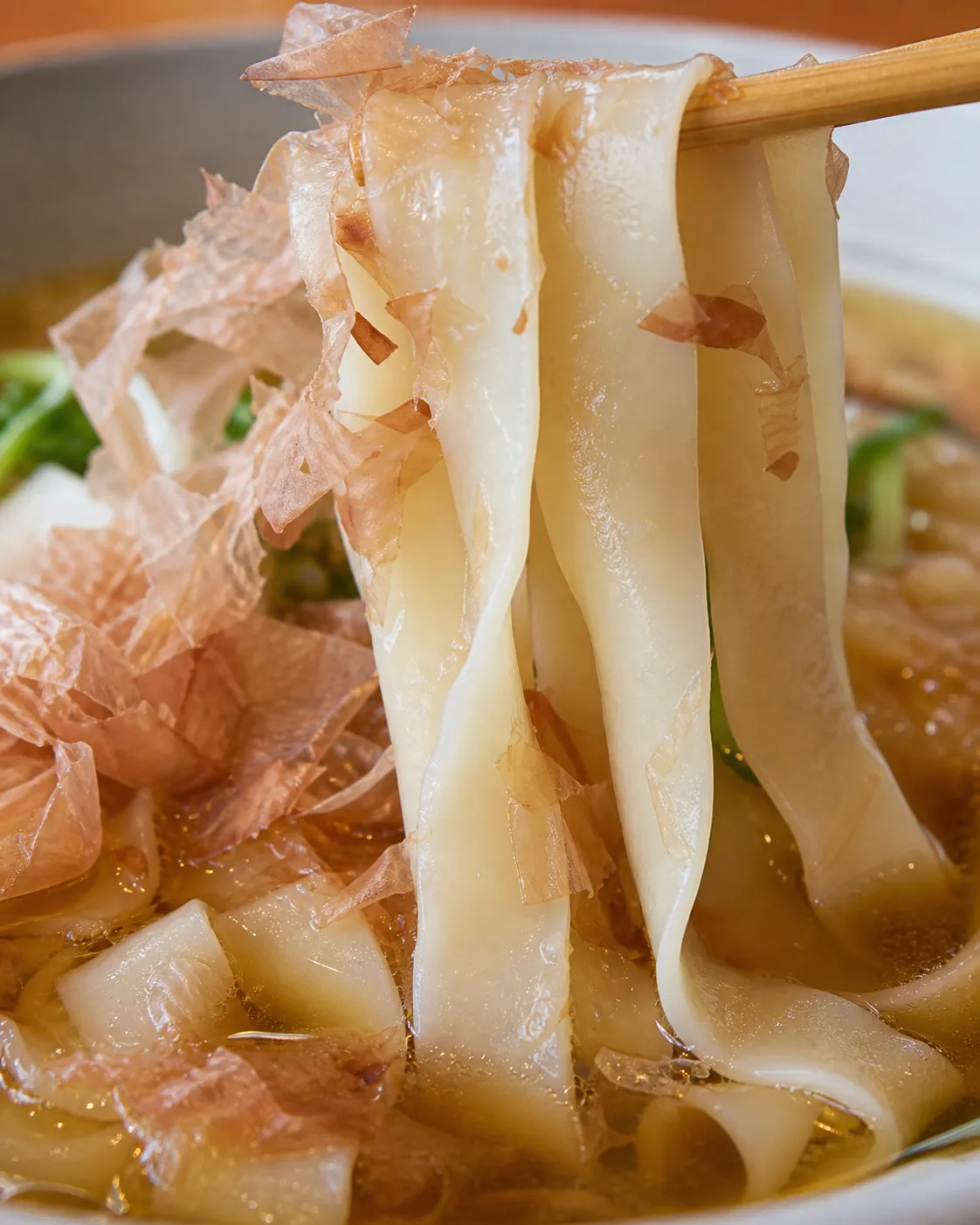 Close-up of flat wide kishimen noodles lifted from a clear light dashi broth with bonito flakes on top