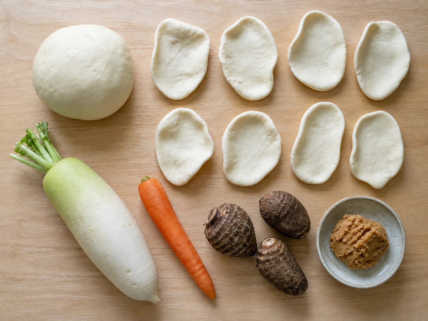 Flat lay of mimi ingredients: wheat dough, hand-pinched ear-shaped pieces, daikon, carrot, taro and miso