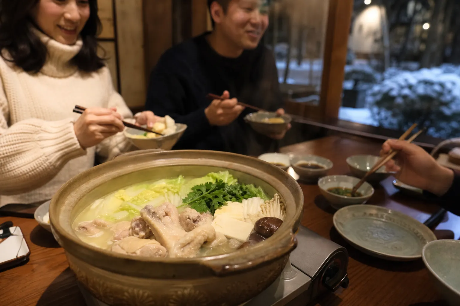 Friends gathered around a bubbling donabe pot of mizutaki chicken hot pot in a cozy Hakata restaurant on a cold night