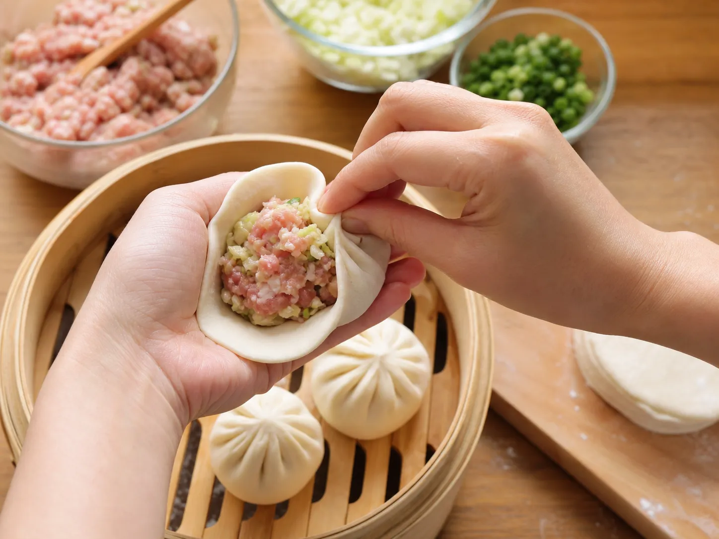 Hands pleating and sealing a nikuman bun over seasoned pork filling on a bamboo steamer