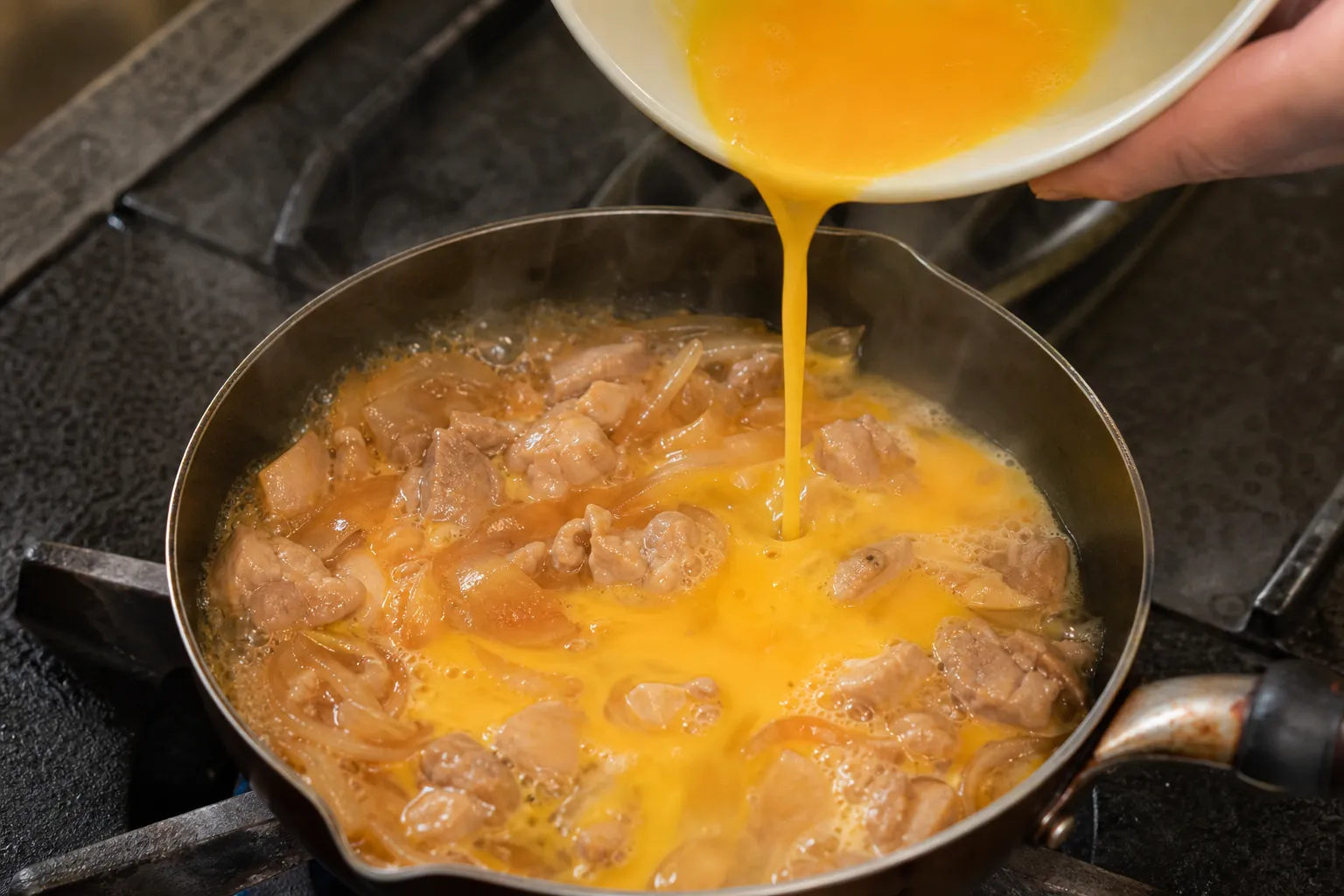 A cook pouring beaten egg over chicken and onion simmering in dashi broth in a shallow oyakodon pan
