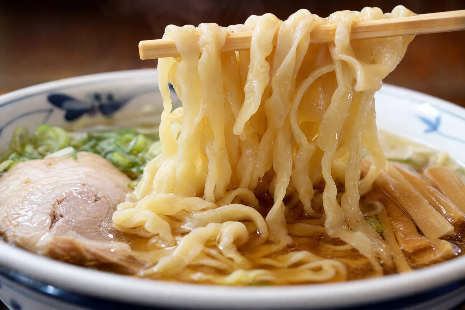 Close-up of flat, wavy hand-pulled Sano ramen noodles lifted from a clear soy broth