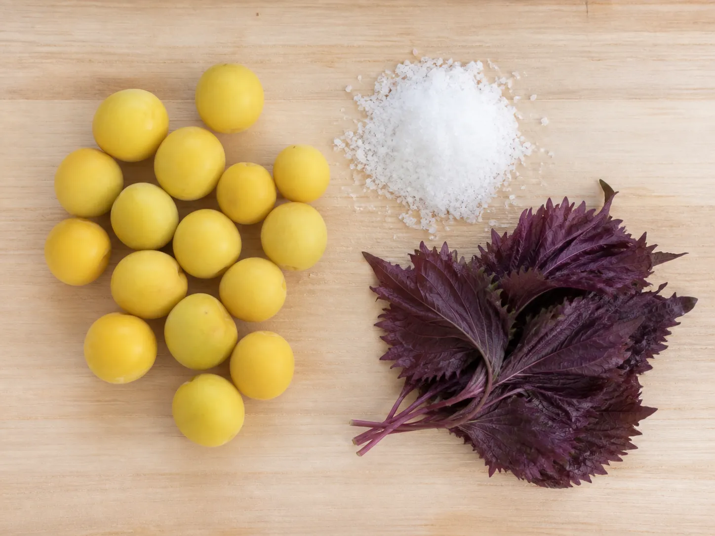 Flat lay of umeboshi ingredients: whole ripe yellow ume, coarse sea salt, and fresh red shiso leaves