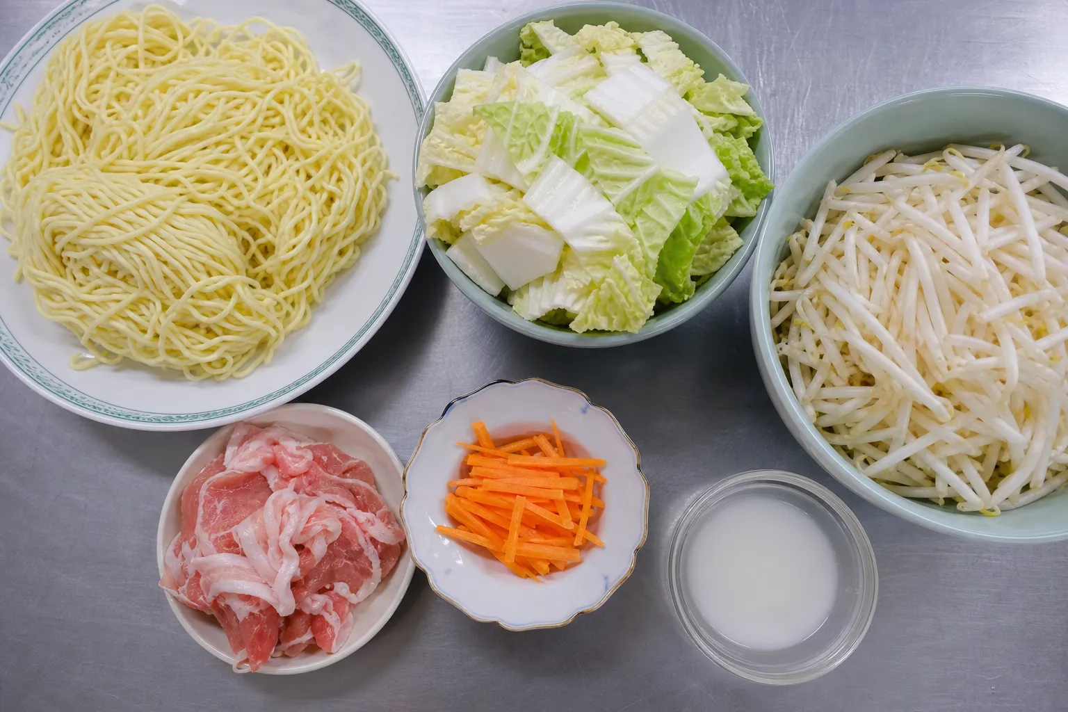 Flat lay of sanma-men ingredients: ramen noodles, napa cabbage, bean sprouts, sliced carrot, thin pork slices and a bowl of starch slurry